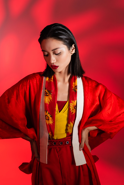 brunette asian woman in neckerchief with floral decor and kimono cape posing with hands on hips on abstract red background