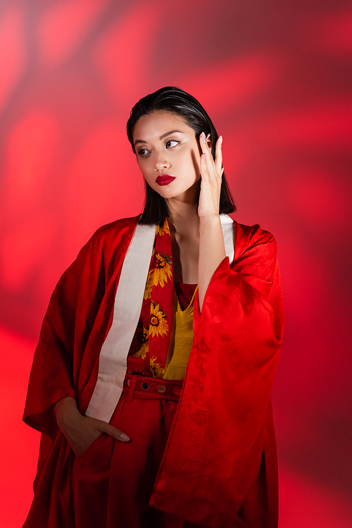 elegant asian woman in kimono cape touching face while standing with hand in pocket on red abstract background