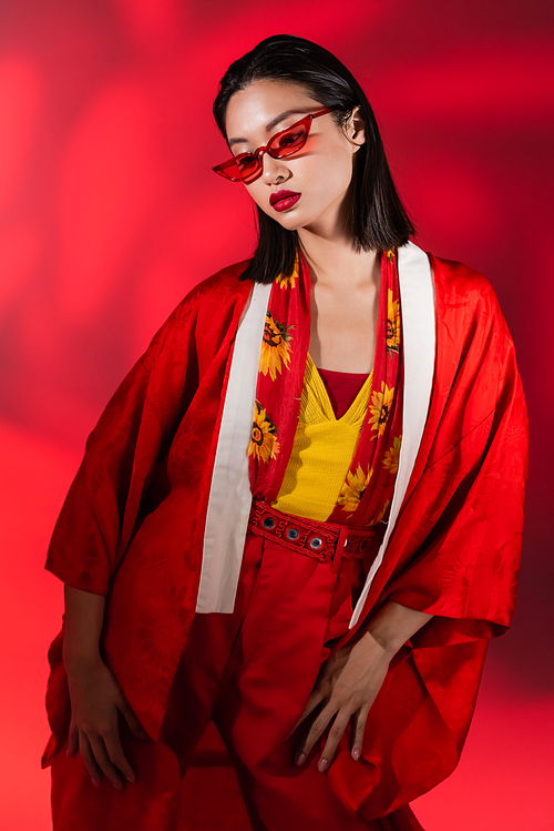 fashionable asian woman in kimono cape and sunglasses posing on red background with shadow