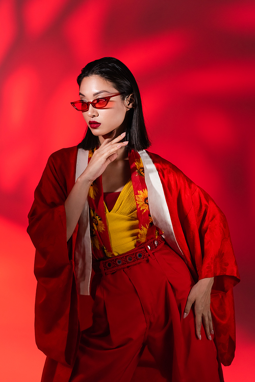 young asian woman in sunglasses and kimono cape posing on abstract background with red light