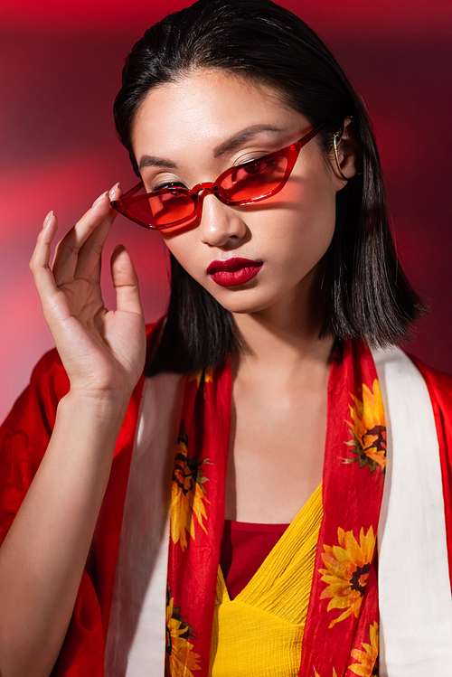 portrait of brunette asian woman touching red sunglasses and looking at camera on dark background