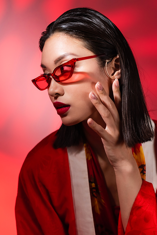 asian woman in trendy sunglasses and ear cuff posing with hand near face on abstract background with red gradient