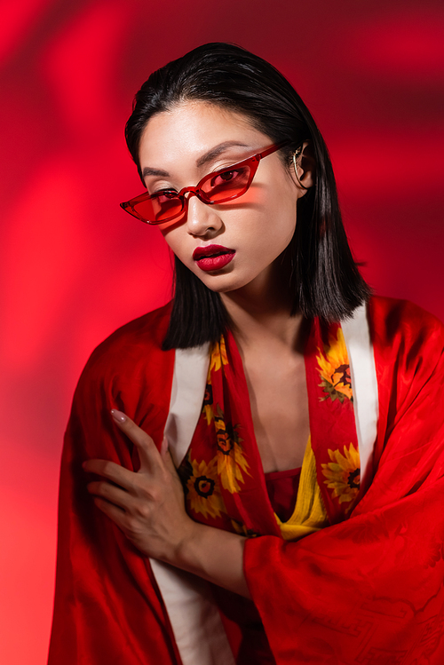 young asian woman in kimono cape and sunglasses looking at camera on abstract red background