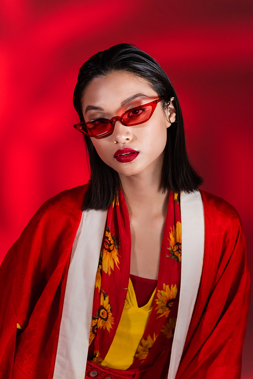 brunette asian woman in elegant kimono cape and stylish sunglasses looking at camera on red background