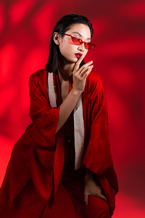 brunette asian woman in kimono cape and sunglasses showing hush sign on red background with shadow