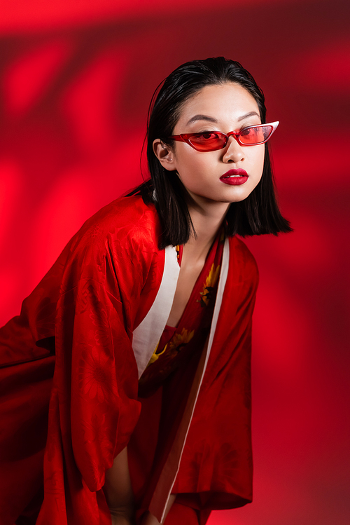 brunette asian woman in stylish sunglasses and kimono cape looking at camera on red  shaded background