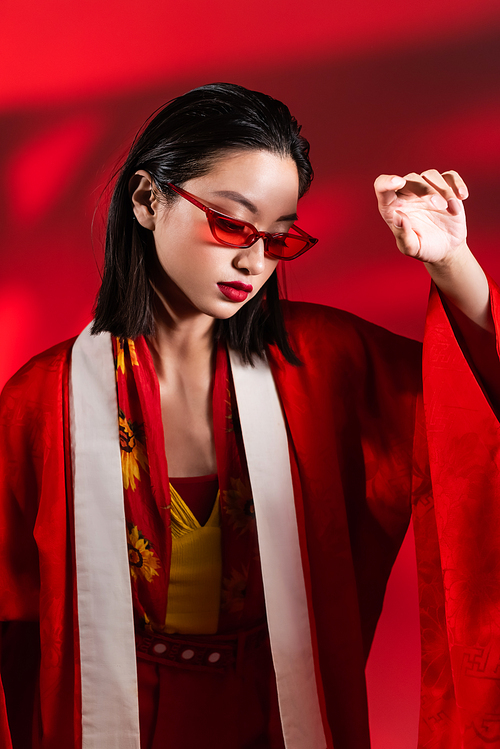 elegant asian woman in stylish sunglasses and kimono cape posing on red background with shadow