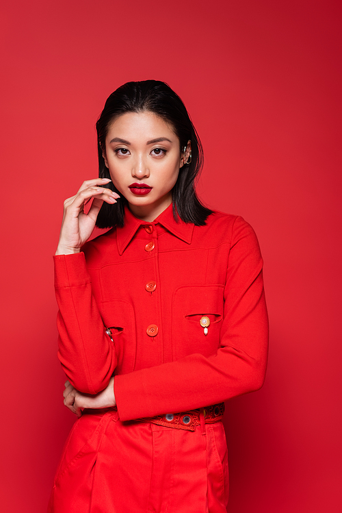 brunette asian woman in fashionable jacket holding hand near face isolated on red