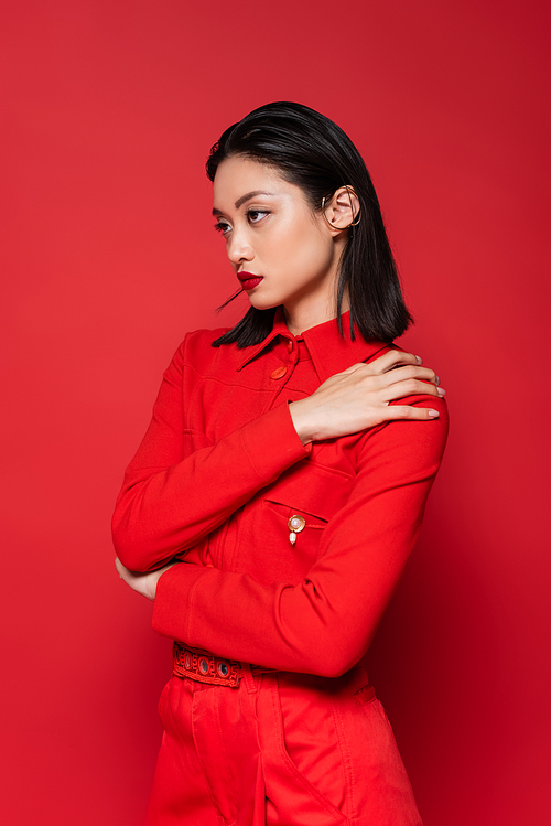 fashionable asian woman in jacket posing with hand on shoulder and looking away isolated on red