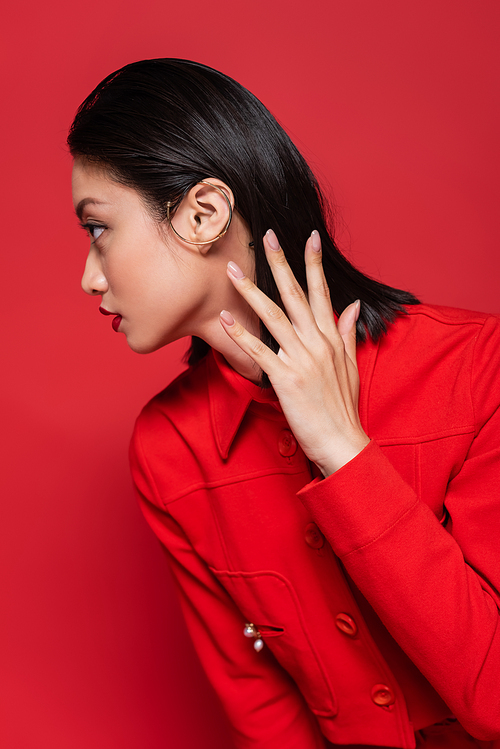 elegant asian woman in jacket and ear cuff holding hand near neck and looking away isolated on red
