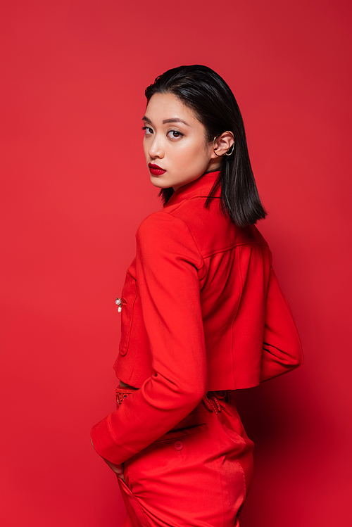 asian woman in stylish clothes standing with hand in pocket and looking at camera on red background