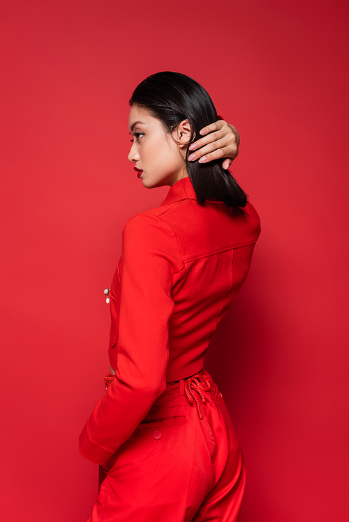 side view of brunette asian woman in stylish suit fixing hair while looking away on red background
