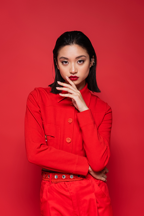 brunette asian woman in fashionable blazer holding hand near face while looking at camera isolated on red