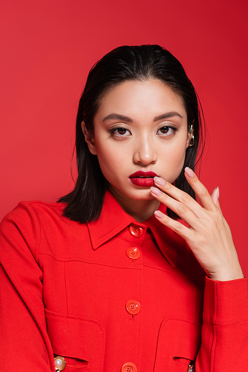 portrait of pretty asian woman in stylish jacket looking at camera and touching lips isolated on red