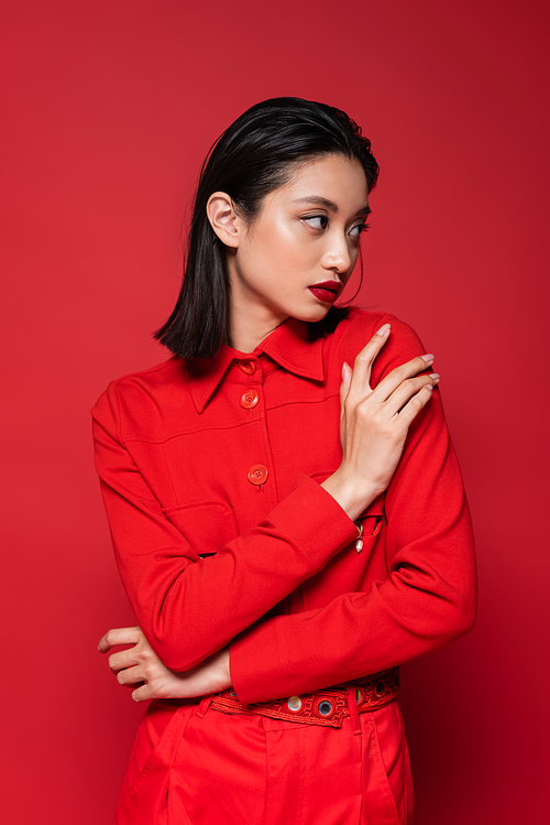 young asian woman with makeup posing in trendy jacket and looking away isolated on red