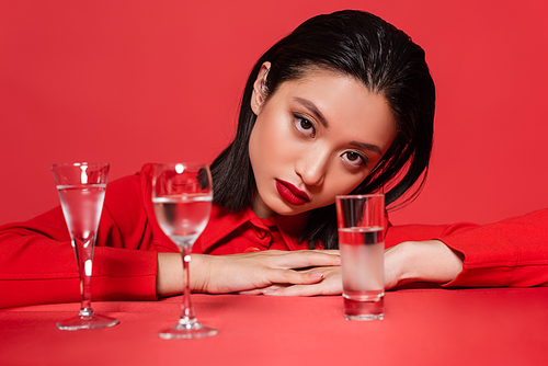 portrait of brunette asian woman in jacket posing near different glasses with water isolated on red