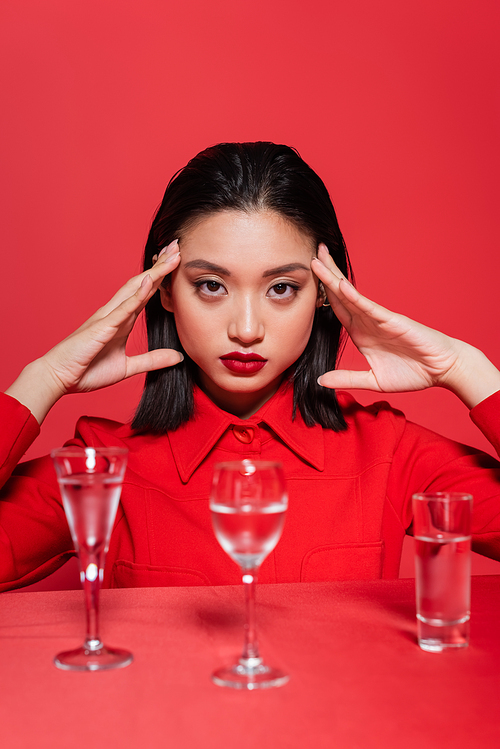 portrait of asian woman in trendy jacket posing near glasses with clear water isolated on red