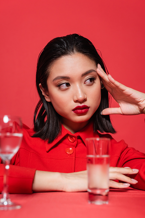 stylish asian woman with makeup holding hand near face and looking away near blurred glasses with water isolated on red
