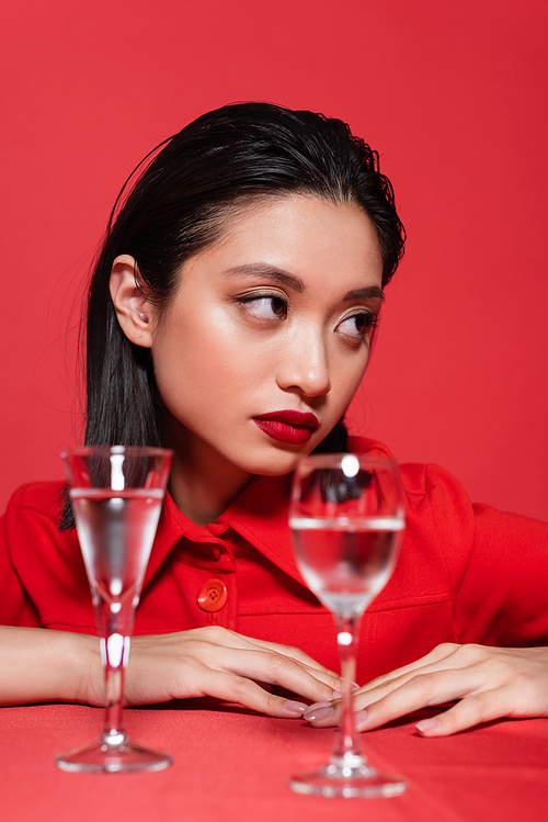 portrait of brunette asian woman with makeup looking away near blurred glasses of water isolated on red