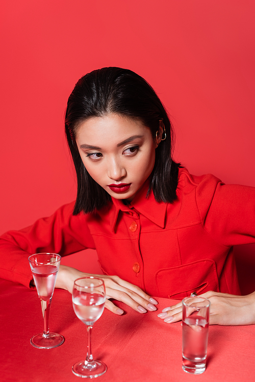 brunette asian woman in stylish jacket looking away near glasses with pure  on red background