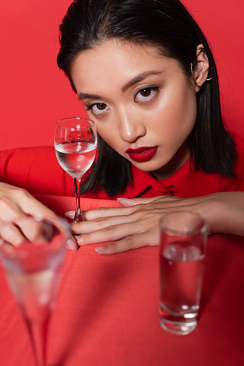 brunette asian woman with makeup and ear cuff looking at camera near glasses with water on blurred foreground isolated on red