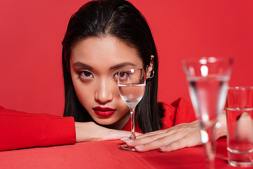 portrait of young asian woman with makeup looking at camera near blurred glasses with water isolated on red