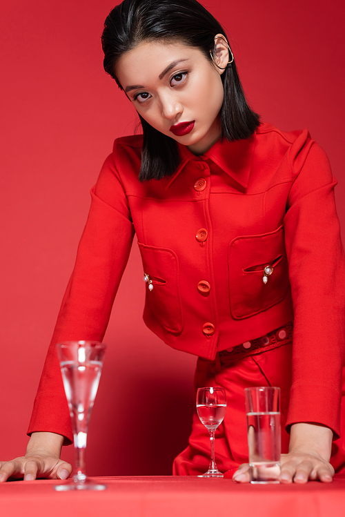 brunette asian woman in elegant jacket standing near glasses with clean  on red background