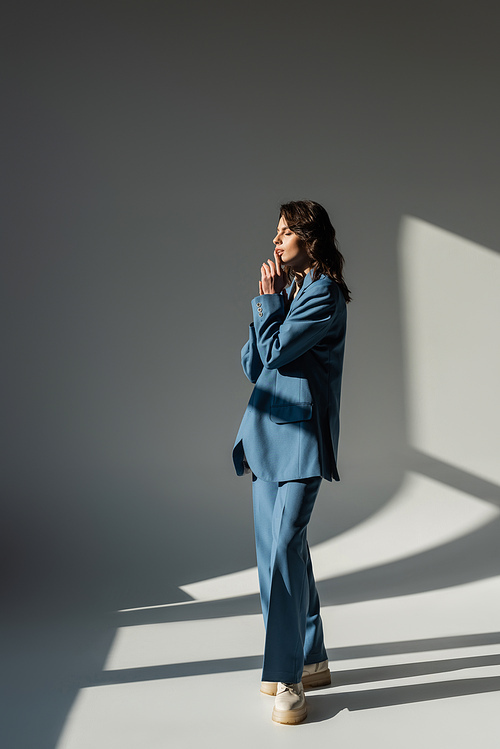 full length of fashionable woman in blue suit standing with closed eyes on grey background with lighting