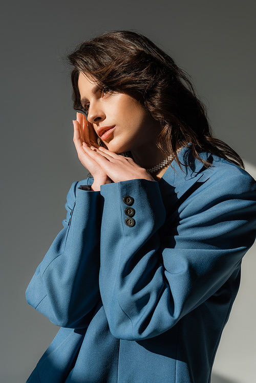 thoughtful brunette woman in blue blazer standing with hands near face and looking away on grey