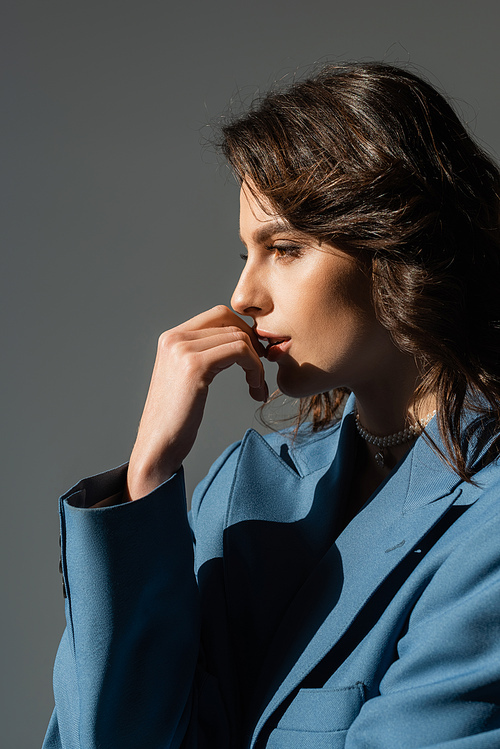 profile of pensive brunette woman in blue blazer holding hand near face isolated on grey