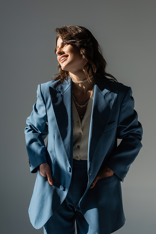 cheerful brunette woman holding hands in pockets of blue and trendy jacket while looking away isolated on grey