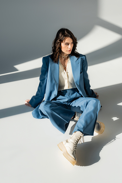 full length of trendy woman in blue suit and white boots sitting on grey background with lighting and shadows