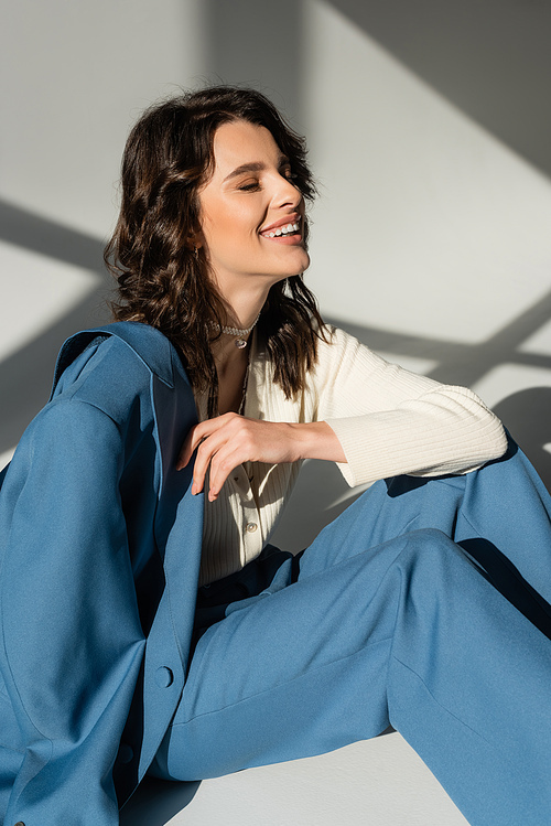 happy brunette woman in blue suit laughing with closed eyes on grey background with lighting and shadows