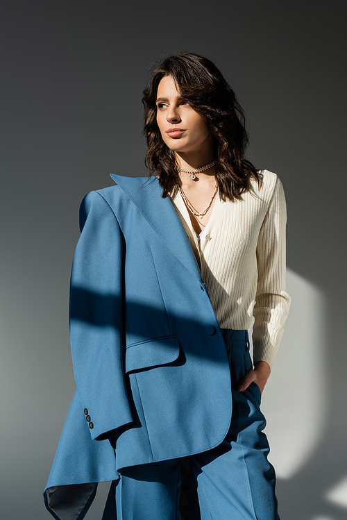 fashionable brunette woman with blue blazer standing with hand in pocket and looking away on grey background