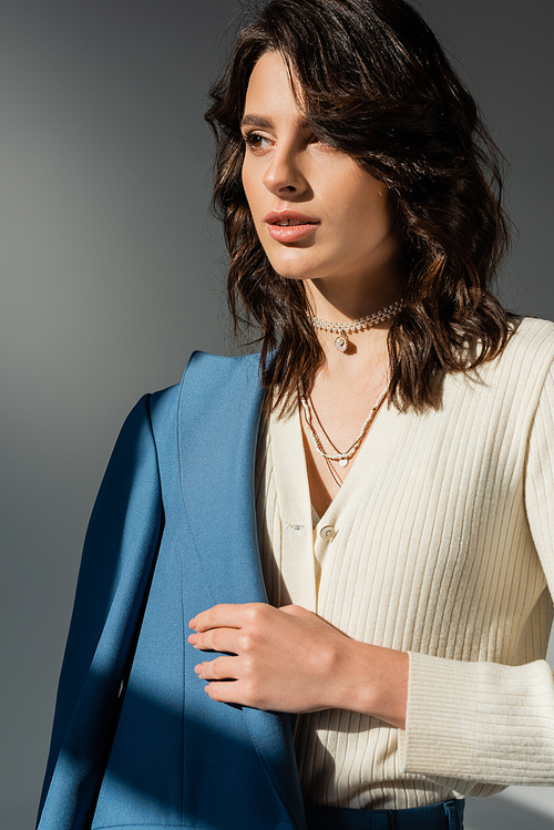 brunette woman in white cardigan and necklaces holding blue blazer and looking away isolated on grey