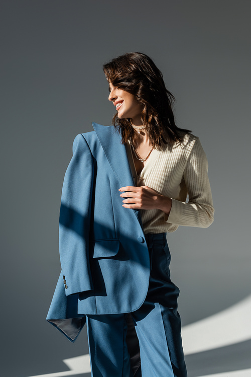happy and stylish woman holding blue blazer while looking away on grey background