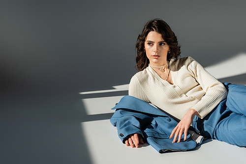 stylish brunette woman looking away while lying near blue jacket on grey background with lighting