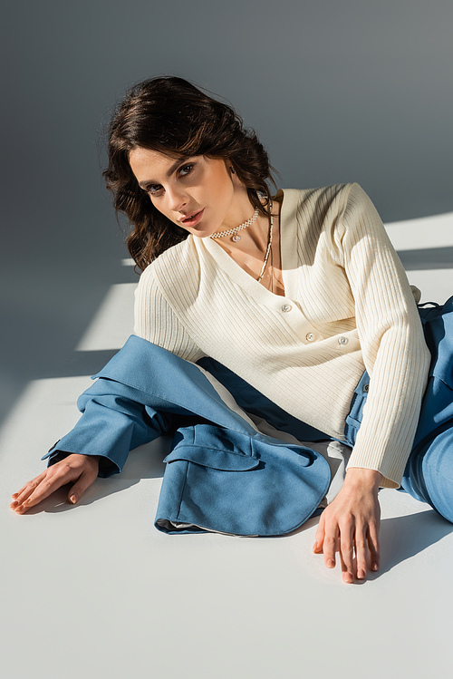 young brunette woman in white jumper looking at camera while lying near blue jacket on grey background with lighting