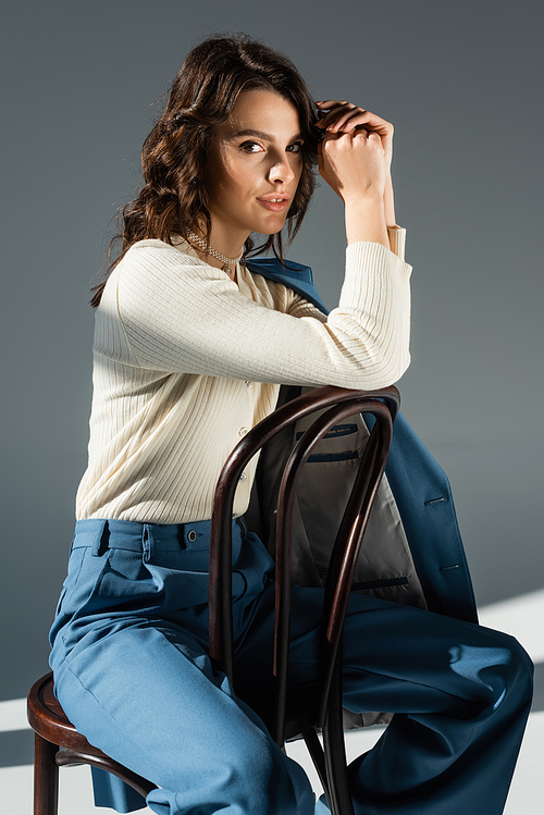 positive woman in white jumper and blue trousers sitting on chair and looking at camera on grey background