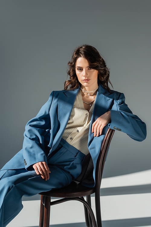 trendy brunette woman in blue oversize suit sitting on chair on grey background with lighting and shadows