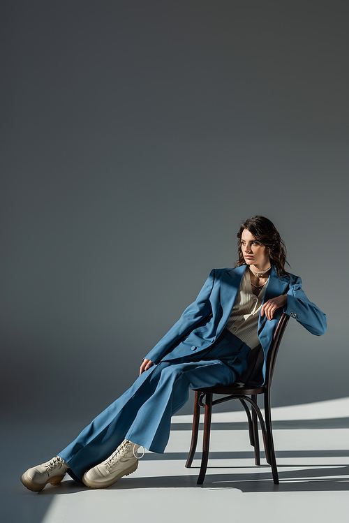 full length of stylish woman in blue suit and boots looking away on chair on grey background with lighting and shadows