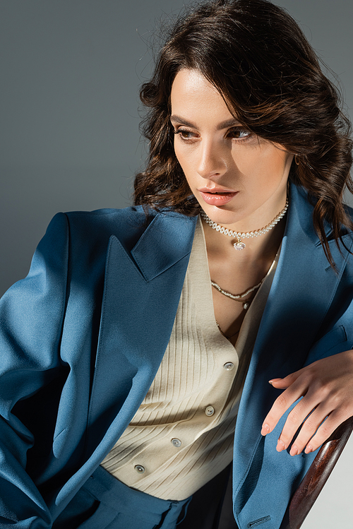 brunette woman in blue blazer and necklaces sitting and looking away on grey background