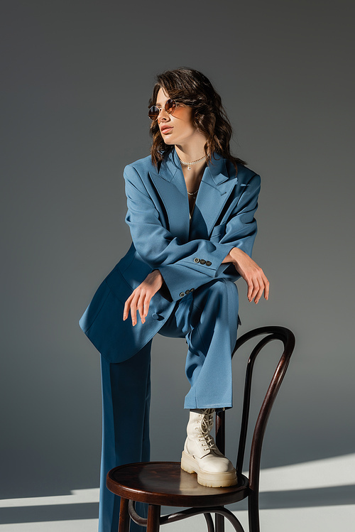 brunette woman in blue fashionable suit stepping on chair and looking away on grey background