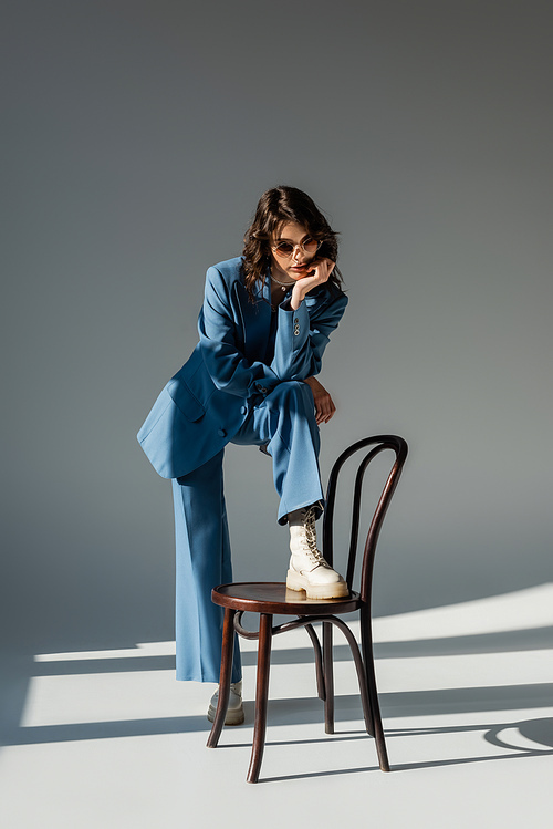 full length of brunette woman in blue suit and trendy sunglasses stepping on chair on grey background with lighting