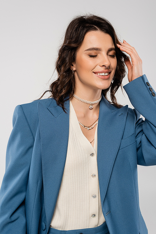 pleased woman in blue blazer and necklaces posing with closed eyes and touching hair isolated on grey