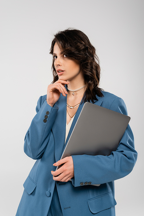 thoughtful brunette woman in blue jacket holding laptop and looking away isolated on grey