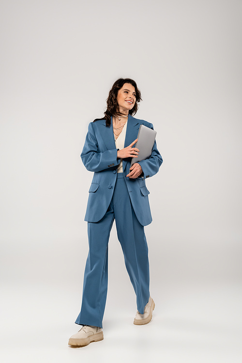 full length of smiling woman in blue suit walking with laptop on grey background