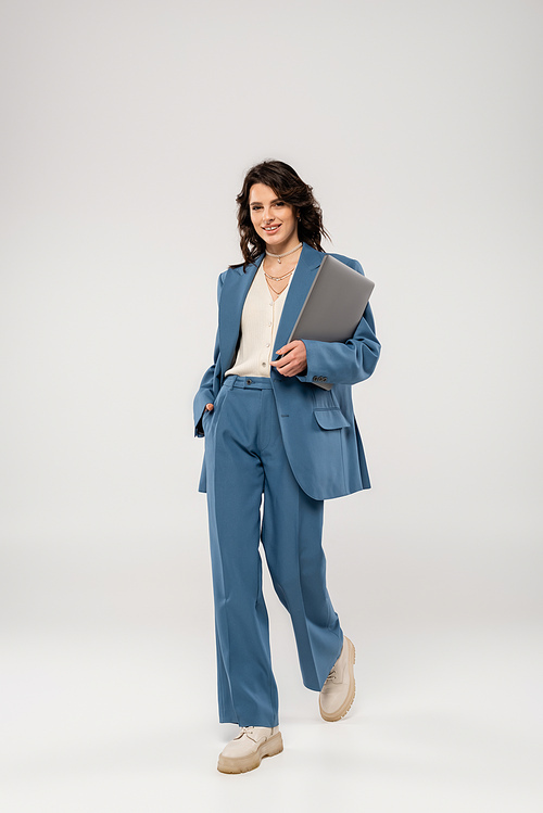 full length of joyful woman in fashionable suit holding hand in pocket while walking with laptop on grey background