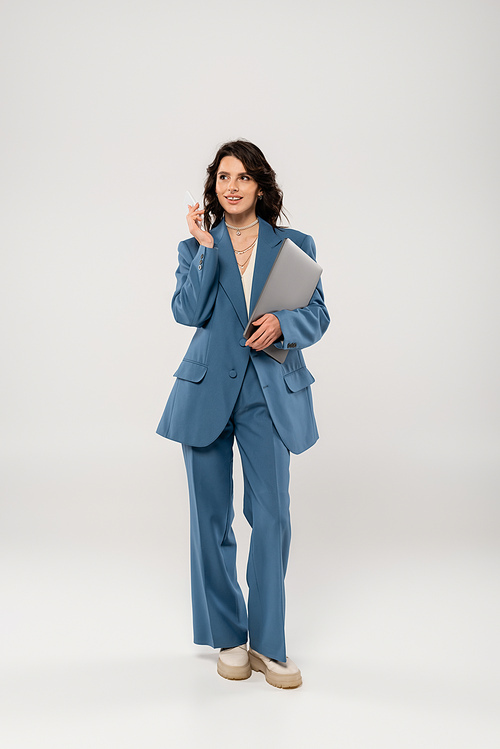 full length of trendy woman in blue suit standing with laptop and mobile phone on grey background