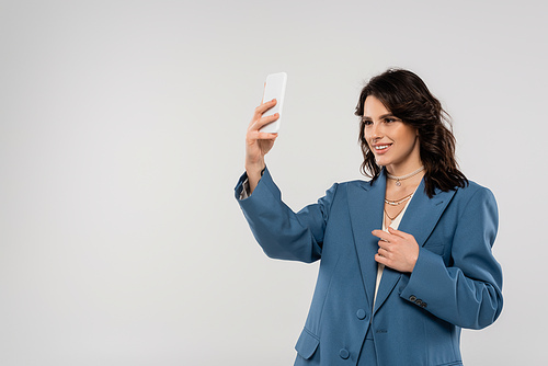 smiling woman in blue blazer and necklaces taking selfie on mobile phone isolated on grey